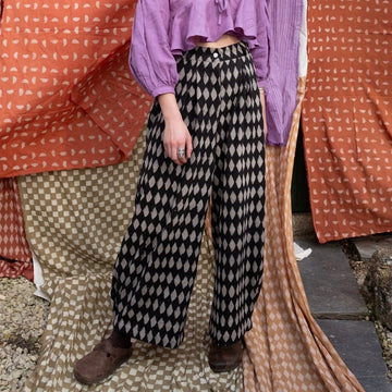 A person standing in front of a backdrop featuring a harlequin print design. Wearing black and beige harlequin print jeans with a purple top.