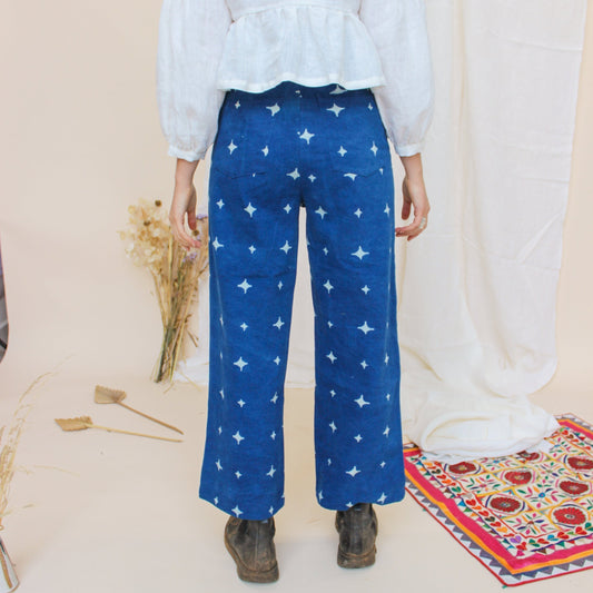 A person standing in a studio setting, wearing high-waisted indigo jeans with a star print, paired with a white top and black boots, in front of a white background with floral props.