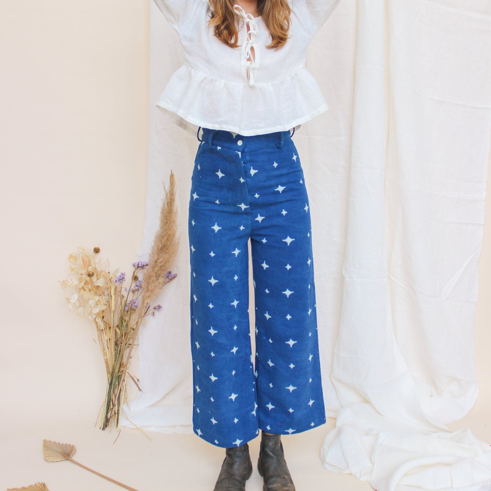 A person standing in a studio setting, wearing high-waisted indigo jeans with a star print, paired with a white top and black boots, in front of a white background with floral props.