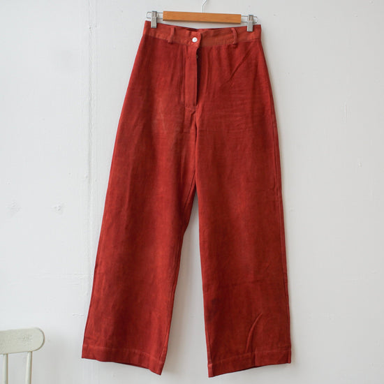A pair of high-waisted, red colored Piper jeans with a button down fastening and two back pockets, hanging on a wooden hanger against a white wall.