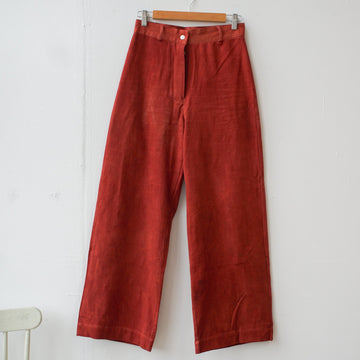 A pair of high-waisted, red colored Piper jeans with a button down fastening and two back pockets, hanging on a wooden hanger against a white wall.