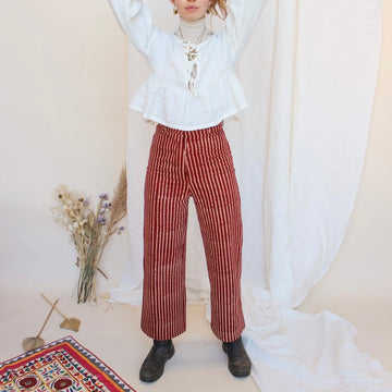 A person standing in front of a white fabric backdrop wearing high-waisted, red and white striped jeans with a white top and black boots. There is a small floral arrangement and a decorative, multicolored textile in the foreground.