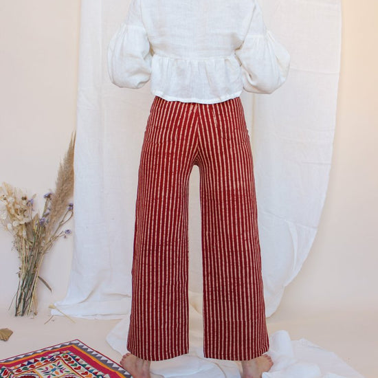 A person standing in front of a white fabric backdrop wearing high-waisted, red and white striped jeans with a white top and black boots. There is a small floral arrangement and a decorative,