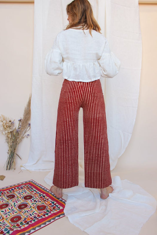 A person standing in front of a white fabric backdrop wearing high-waisted, red and white striped jeans with a white top and black boots. There is a small floral arrangement and a decorative,