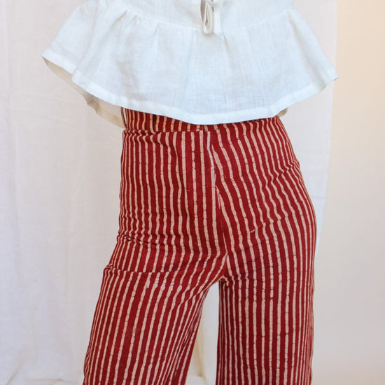 A person standing in front of a white fabric backdrop wearing high-waisted, red and white striped jeans with a white top and black boots. There is a small floral arrangement and a decorative,