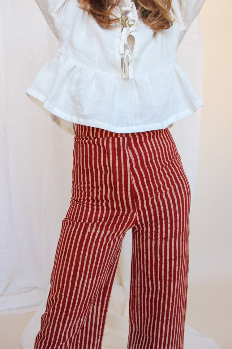 A person standing in front of a white fabric backdrop wearing high-waisted, red and white striped jeans with a white top and black boots. There is a small floral arrangement and a decorative,