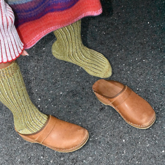 A woman wearing a coloured skirt, green socks and brown clogs on a grey floor.