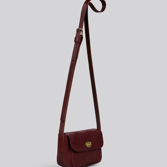 Maroon red  leather crossbody bag with brass clasp on a light gray background