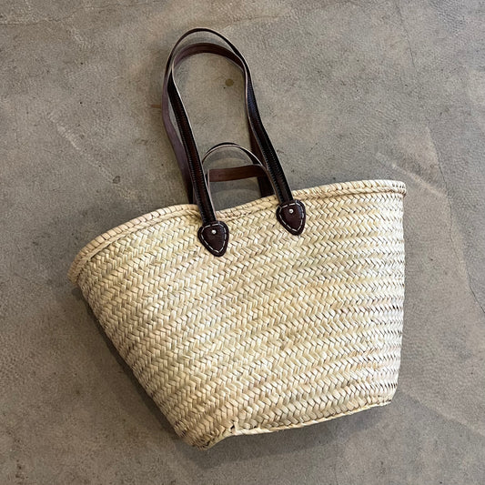 A beige woven basket with a herringbone pattern and double straps made of dark leather, placed on a grey surface.