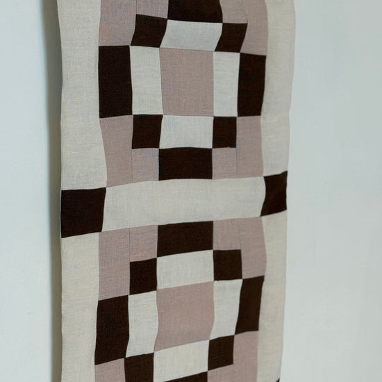 Checkered fabric wall hanging with brown, beige, and black squares on a white background