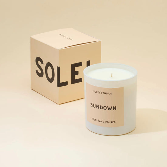A white candle labeled 'Sundown' by Solei Studios, next to its packaging box with the word 'SOLEI' on it, all set against a beige background.