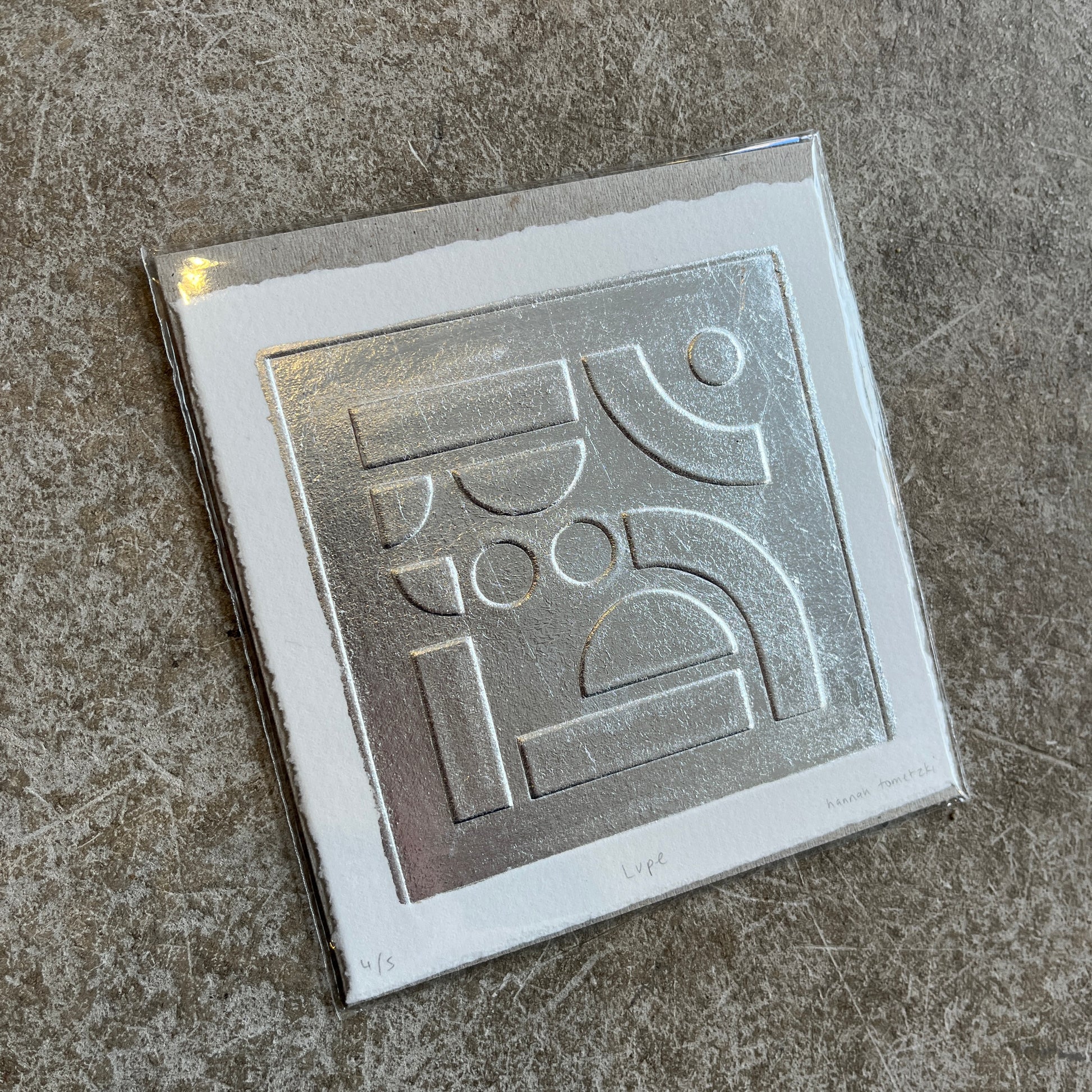 Silver leaf embossed geometric abstract print with a white boarder on heritage cotton paper. Square shaped on a grey concrete background