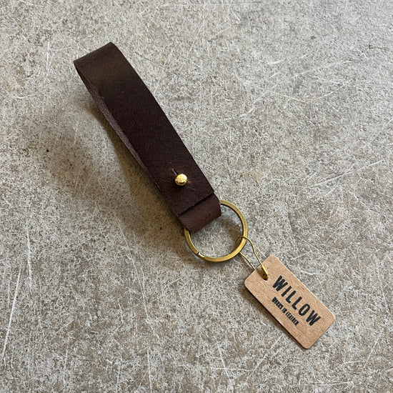 A collection of leather loop keyrings in various colors including red, navy, green, brown, and beige, with solid brass details and branded tags.