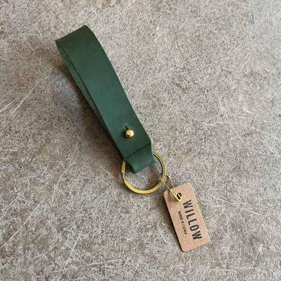 A collection of leather loop keyrings in various colors including red, navy, green, brown, and beige, with solid brass details and branded tags.
