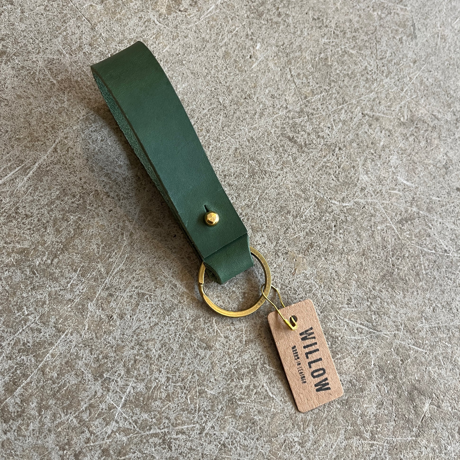 A collection of leather loop keyrings in various colors including red, navy, green, brown, and beige, with solid brass details and branded tags.