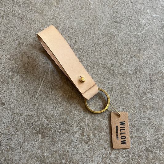 A collection of leather loop keyrings in various colors including red, navy, green, brown, and beige, with solid brass details and branded tags.