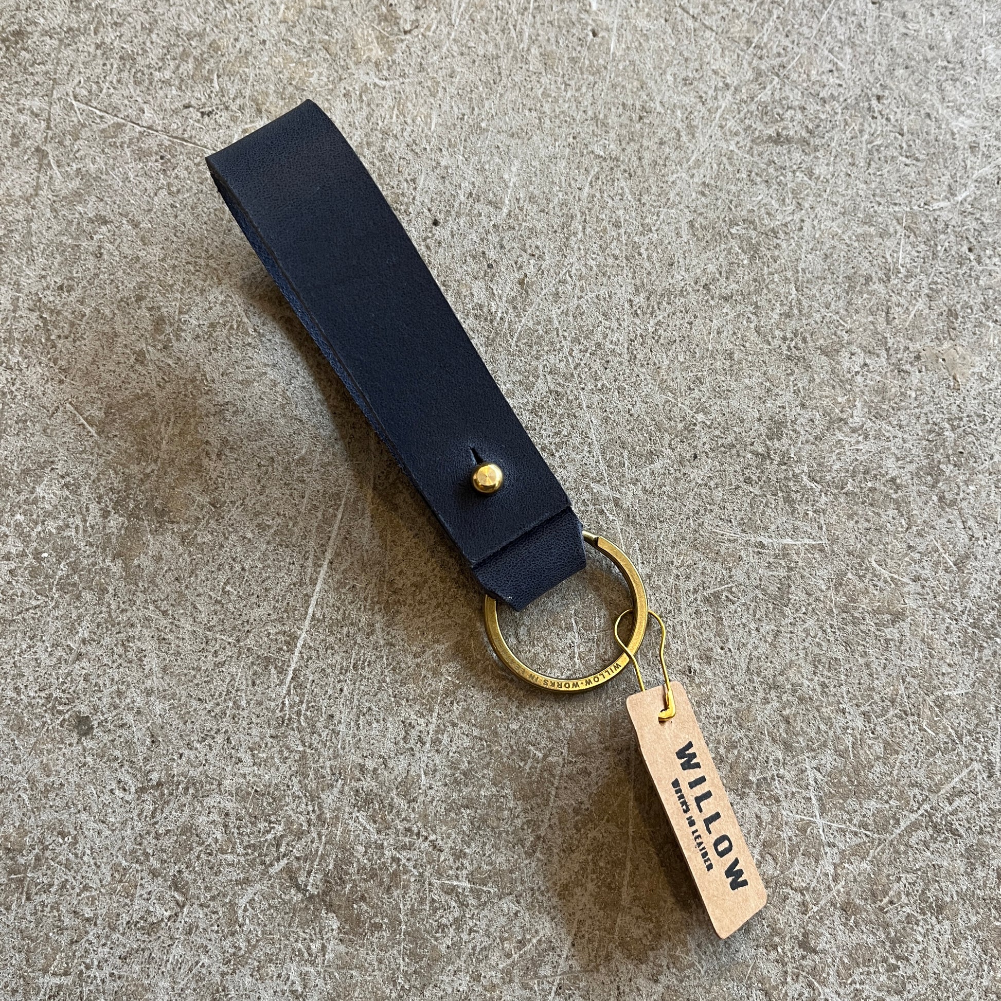 A collection of leather loop keyrings in various colors including red, navy, green, brown, and beige, with solid brass details and branded tags.