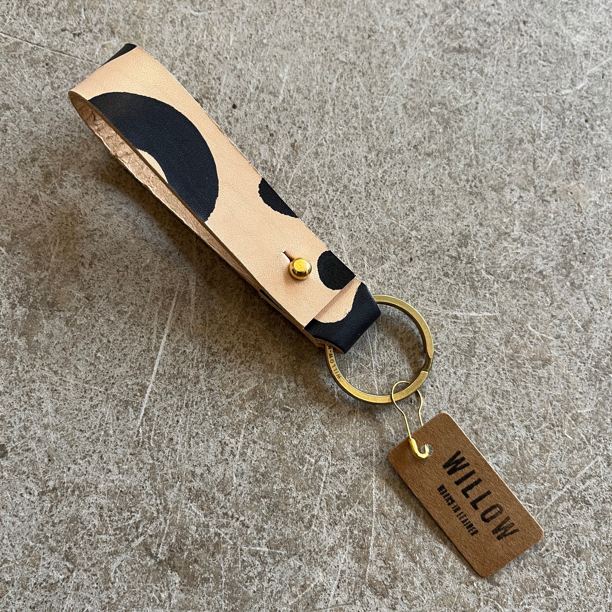 A collection of leather loop keyrings in various colors including red, navy, green, brown, and beige, with solid brass details and branded tags.