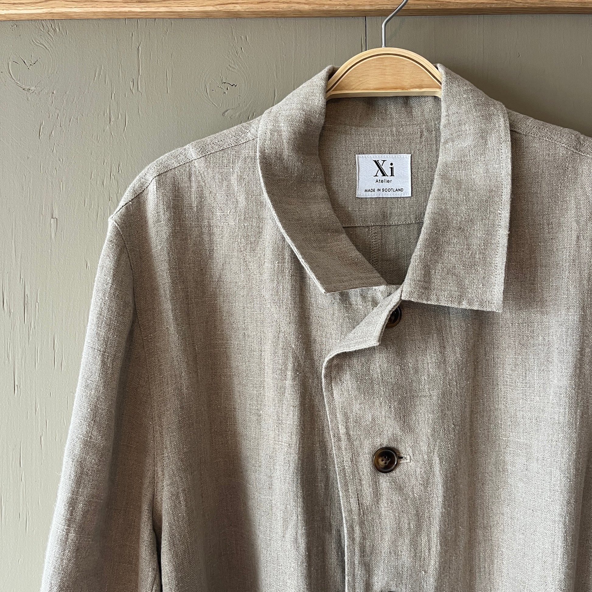 Beige linen coat on a hanger with 'Xi' label against a neutral background