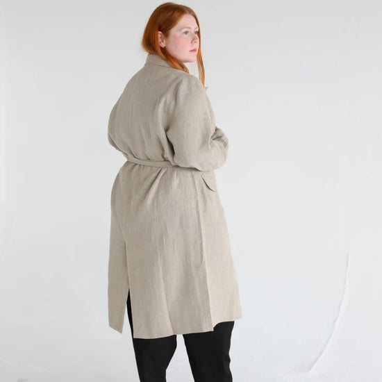 A person viewed from the back wearing a beige linen coat with a belt, standing in a studio setting.