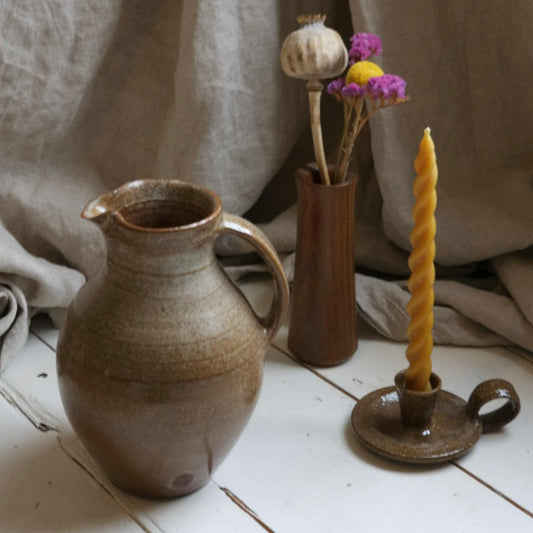 Ceramic pitcher, vase with dried flowers, and candle holder on a wooden surface with a beige fabric background.