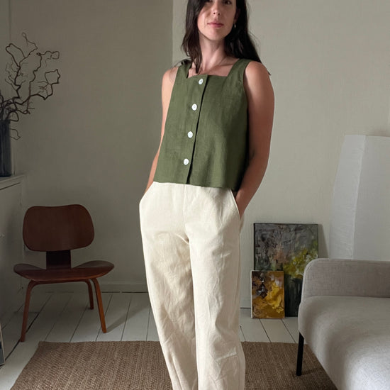Woman wearing a green sleeveless top and white pants in a living room setting.