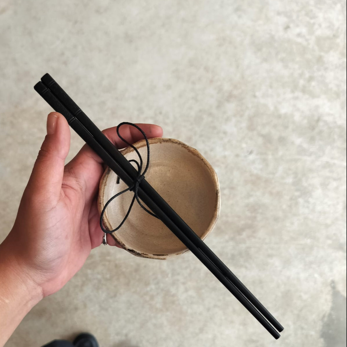 Hand holding black chopsticks over a beige ceramic bowl on a textured surface