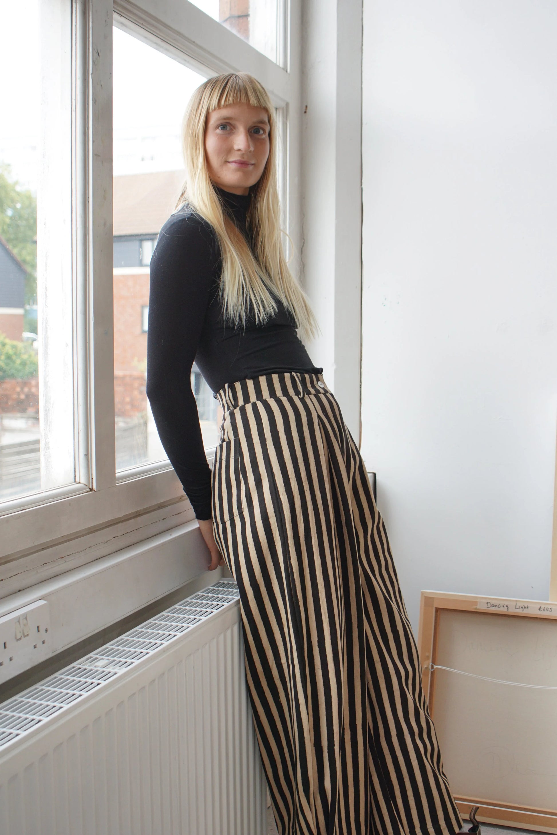 A woman leaning by a window wearing a black top and striped black and white trousers