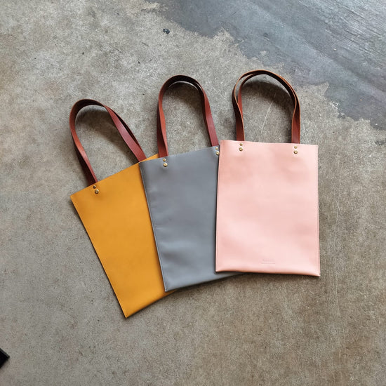Three tote bags in yellow, gray, and pink on a concrete surface