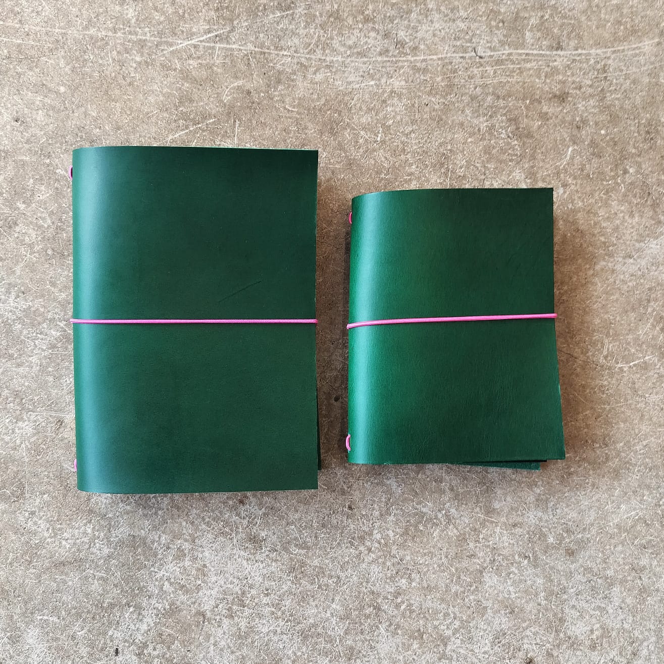 Two green leather-bound books with pink elastic bands on a textured surface