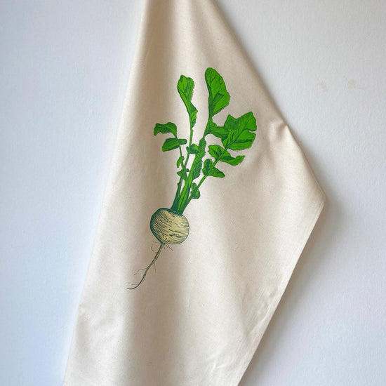 A beige cotton tea towel with a printed design of a single turnip with green leaves.