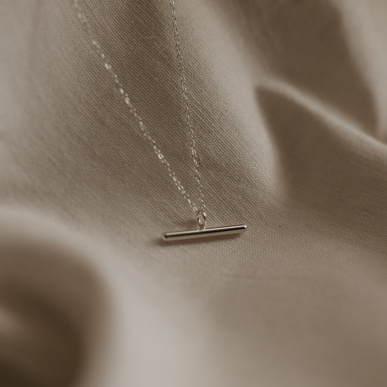 A silver bar necklace with a high polish pendant on a delicate trace chain, displayed on a textured background.