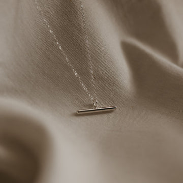 A silver bar necklace with a high polish pendant on a delicate trace chain, displayed on a textured background.