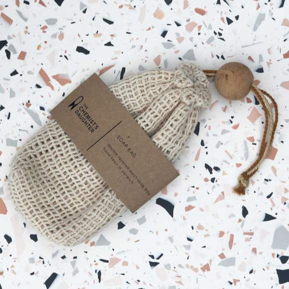 A beige cotton mesh soap bag with a drawstring and a cork ball, branded with 'The Chemist's Daughter' on a brown paper tag.