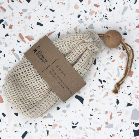 A beige cotton mesh soap bag with a drawstring and a cork ball, branded with 'The Chemist's Daughter' on a brown paper tag.