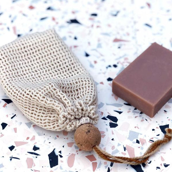 A beige cotton mesh soap bag with a drawstring and a cork ball, branded with 'The Chemist's Daughter' on a brown paper tag.