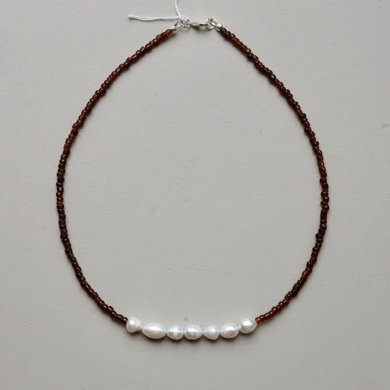 A beaded necklace featuring dark brown seed beads with a horizontal row of five white freshwater pearls across the center, finished with a silver clasp.