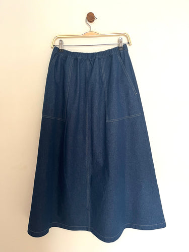 Indigo denim aline midi skirt with an elasticated waist band and utility side pockets on a cream background