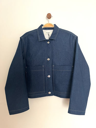 Indigo denim boxy cropped workers jacket with a collar, long sleeves, silver front buttons and patch pockets on a cream background