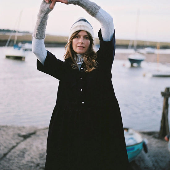 Person in a long navy cord dress with arm arm warmers on and knit hat stretching outdoors by a body of water with boats
