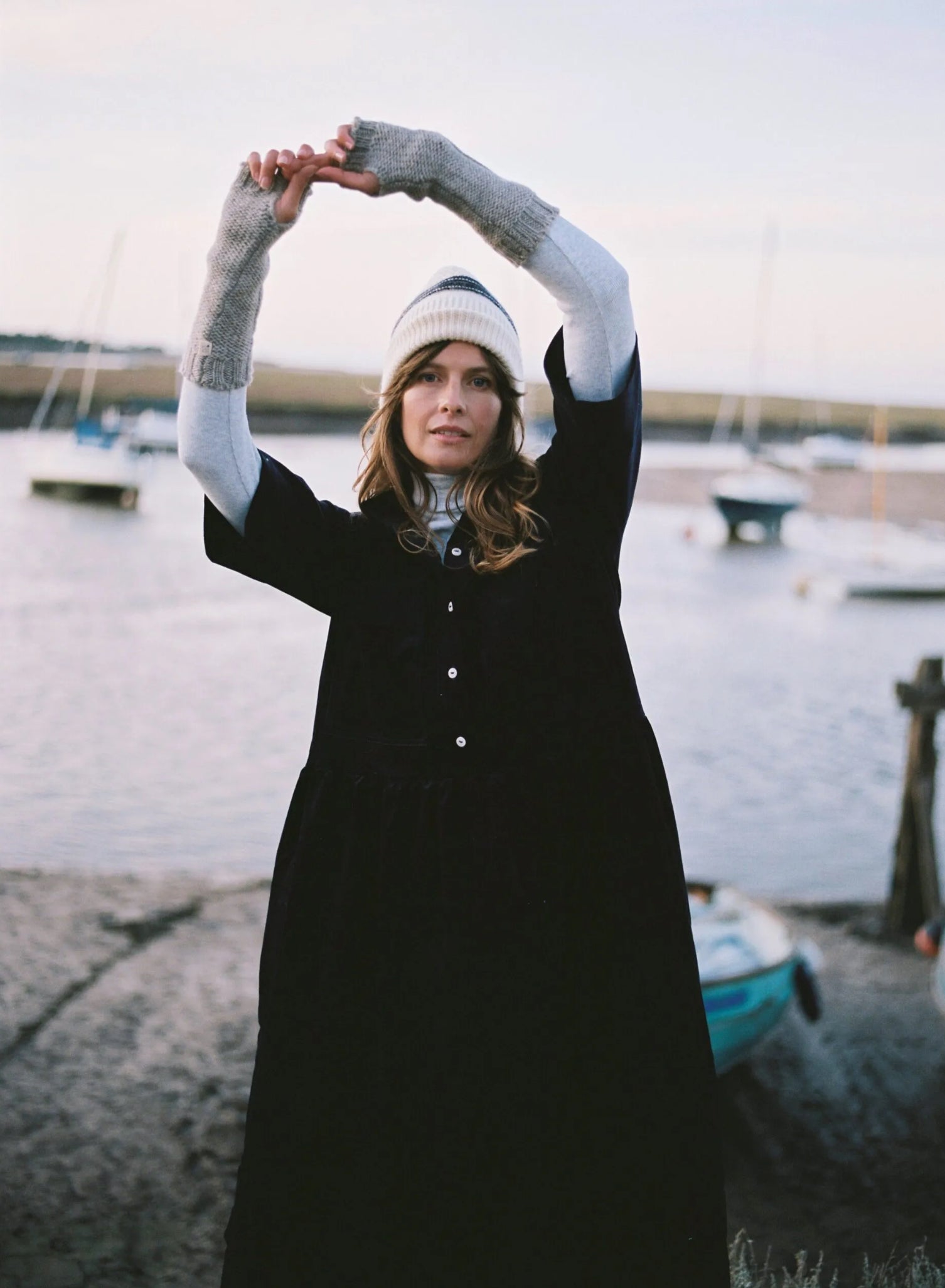 Person in a long navy cord dress with arm arm warmers on and knit hat stretching outdoors by a body of water with boats