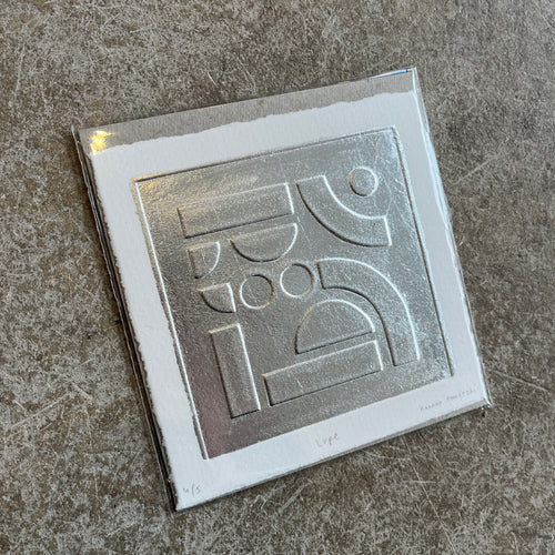 Silver leaf embossed geometric abstract print with a white boarder on heritage cotton paper. Square shaped on a grey concrete background