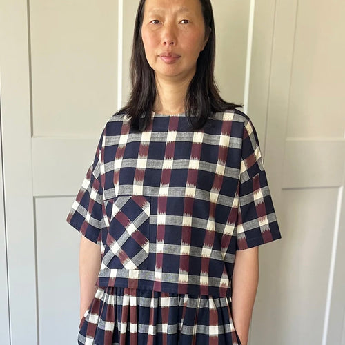 Ikat check in navy blue, brown and cream boxy square fit midi length sleeve top with patch front pocket above hem to one side. Modelled on female with mid length dark hair standing against a white panelled background
