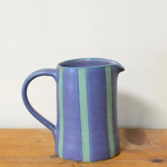 A small ceramic pitcher with a blue body and vertical stripes in light green and darker purple, sitting on a wooden surface against a plain off-white wall.