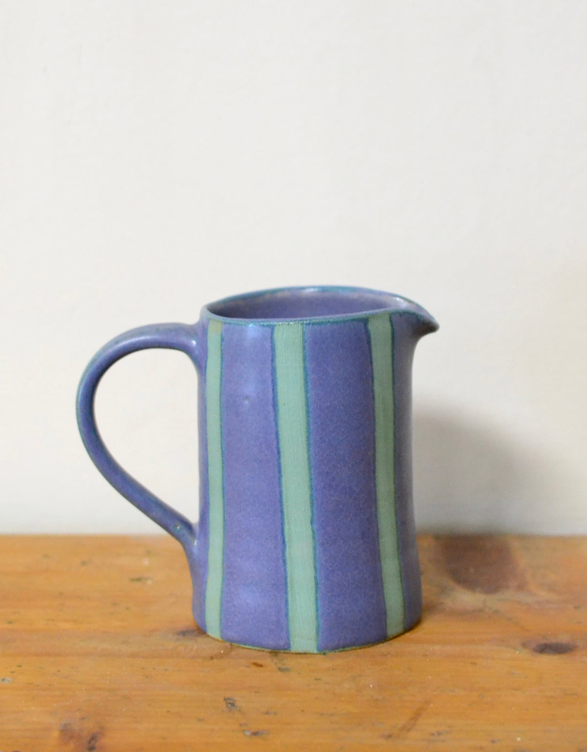 A small ceramic pitcher with a blue body and vertical stripes in light green and darker purple, sitting on a wooden surface against a plain off-white wall.