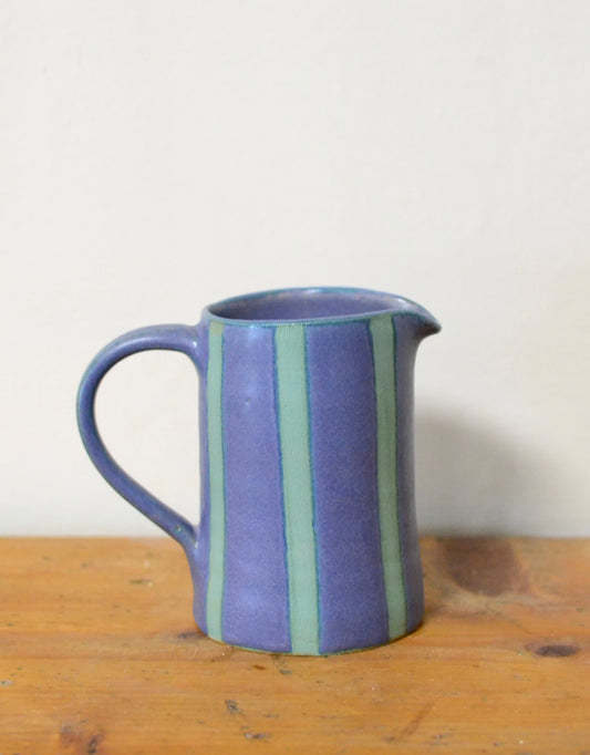 A small ceramic pitcher with a blue body and vertical stripes in light green and darker purple, sitting on a wooden surface against a plain off-white wall.