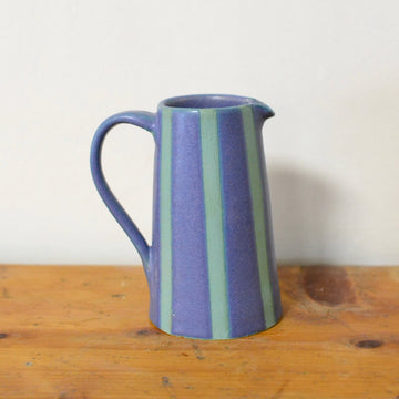 A small ceramic pitcher with a blue body and vertical sage green stripes, sitting on a rustic wooden surface against a plain off-white wall.