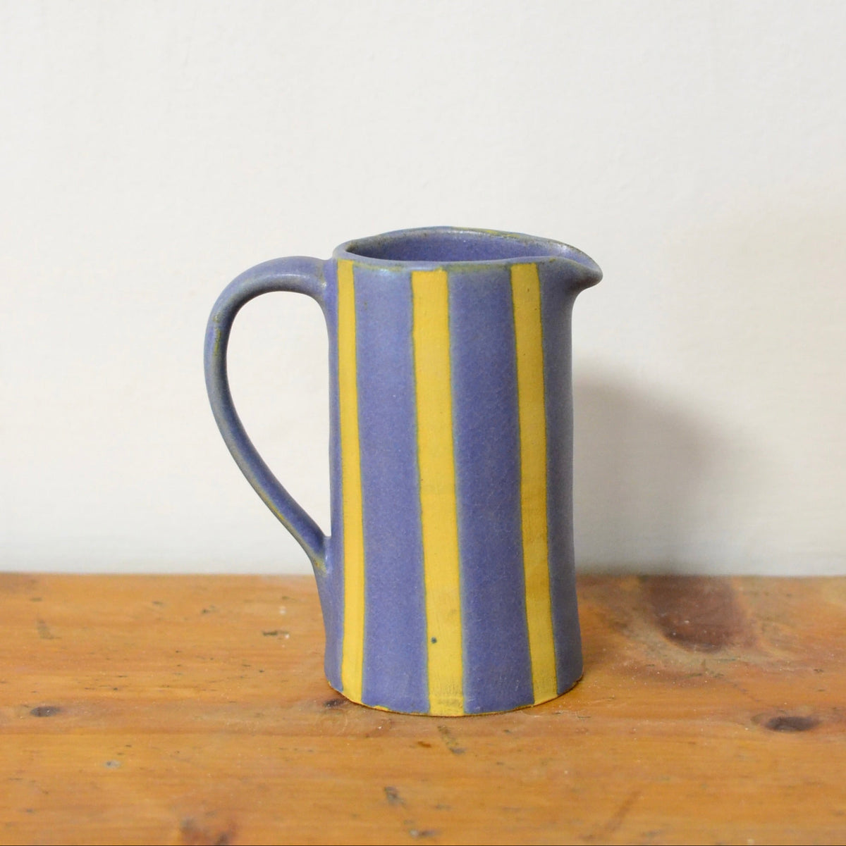 A small ceramic pitcher with a handle, featuring vertical yellow stripes on a muted blue glaze, sitting on a rustic wooden surface against a plain off-white wall.