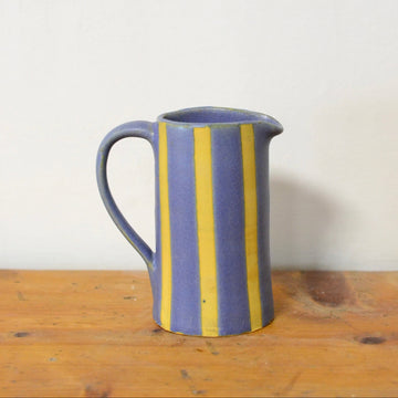 A small ceramic pitcher with a handle, featuring vertical yellow stripes on a muted blue glaze, sitting on a rustic wooden surface against a plain off-white wall.