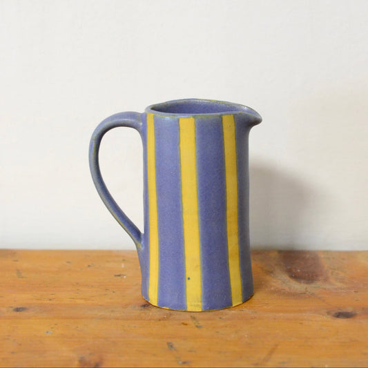 A small ceramic pitcher with a handle, featuring vertical yellow stripes on a muted blue glaze, sitting on a rustic wooden surface against a plain off-white wall.